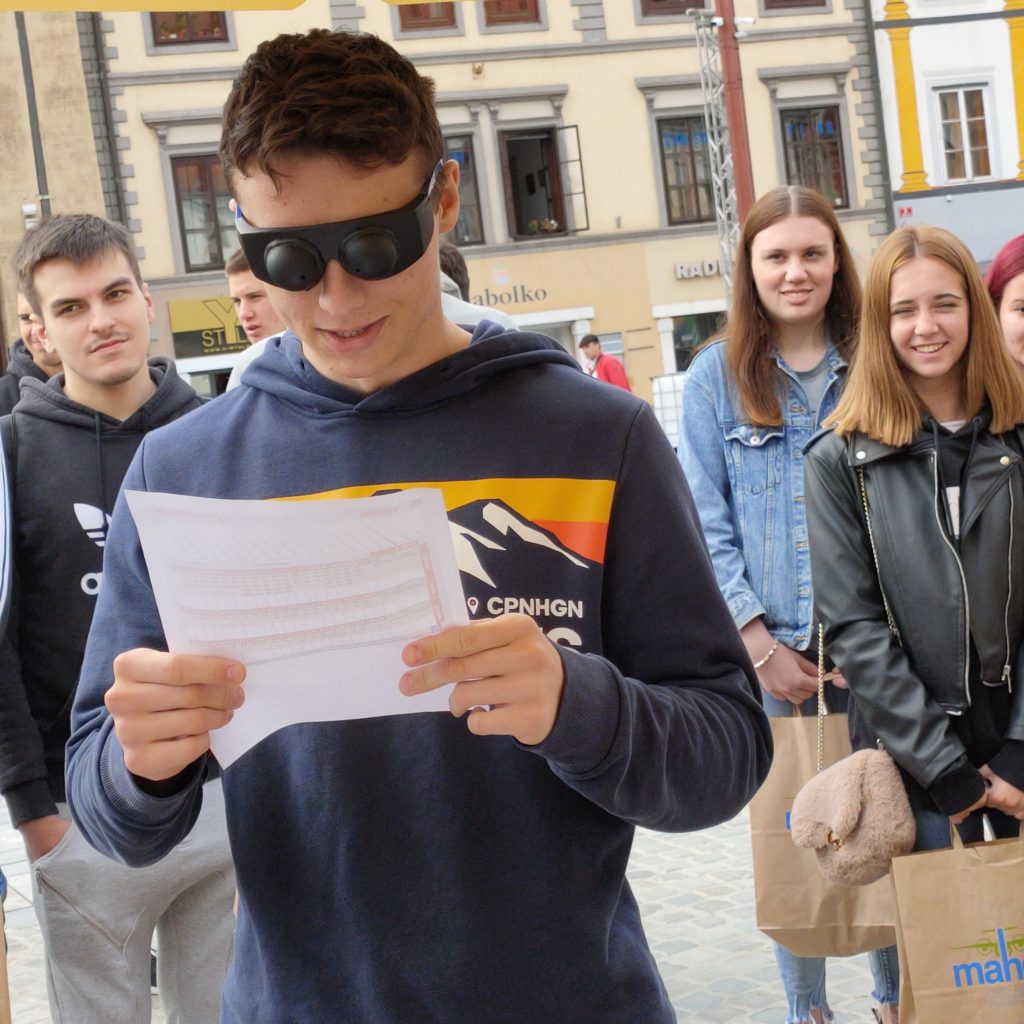 Dijak poskuša s simulacijskimi očali za tunelski vid brati avtobusni vozni red.