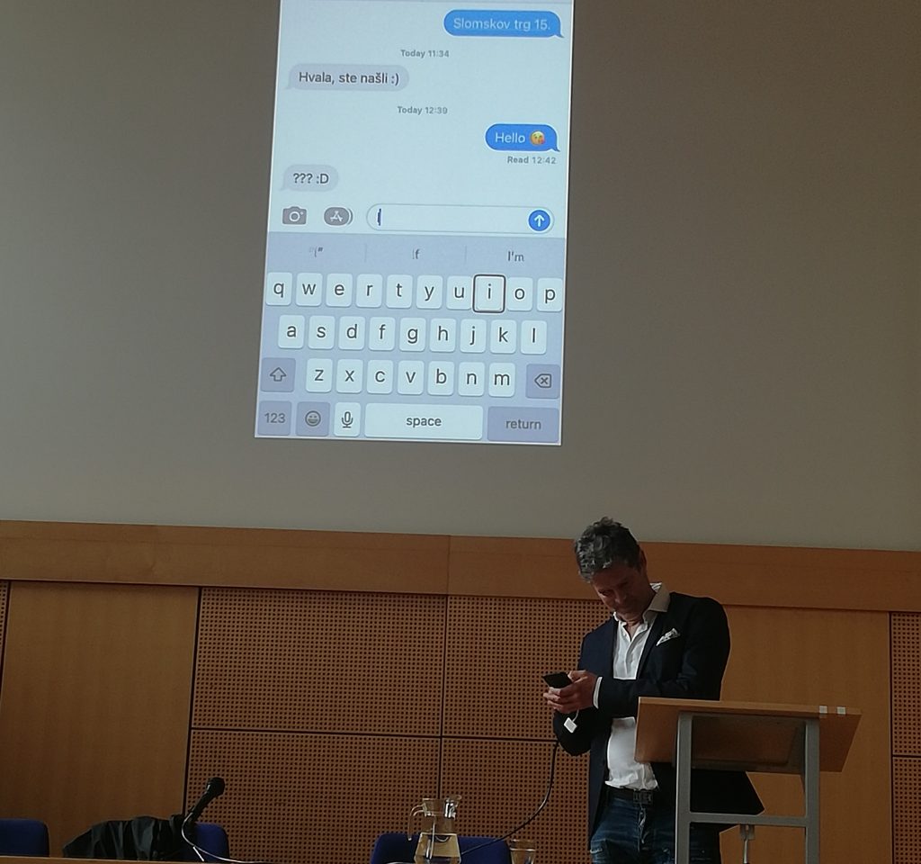 Alen Kobilica stoji s telefonom v roki na govorniškem odru Univerze v Mariboru.