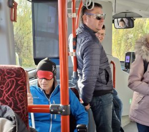 Udeleženec z belo palico in prekritimi očmi sedi na avtobusu, drug pa s simulacijskimi očali stoji na avtobusu