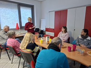 Manja stoje predstavlja svoj izkušnjo, udeleženci pa sedijo za dolgo mizo in poslušajo