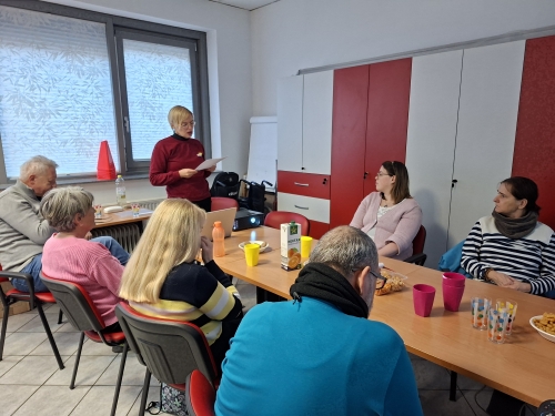 Manja stoje predstavlja svoj izkušnjo, udeleženci pa sedijo za dolgo mizo in poslušajo