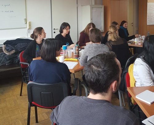 Udeleženci sedijo za štirimi pravokotnimi mizami in poslušajo predavateljico