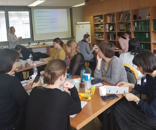 Udeleženci sedijo za štirimi mizami in poslušajo predavateljico