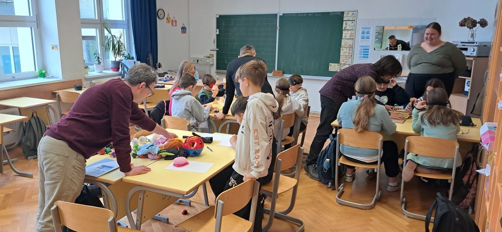 Za več mizami učenci z različnimi pripomočki spoznavajo posebne potrebe.